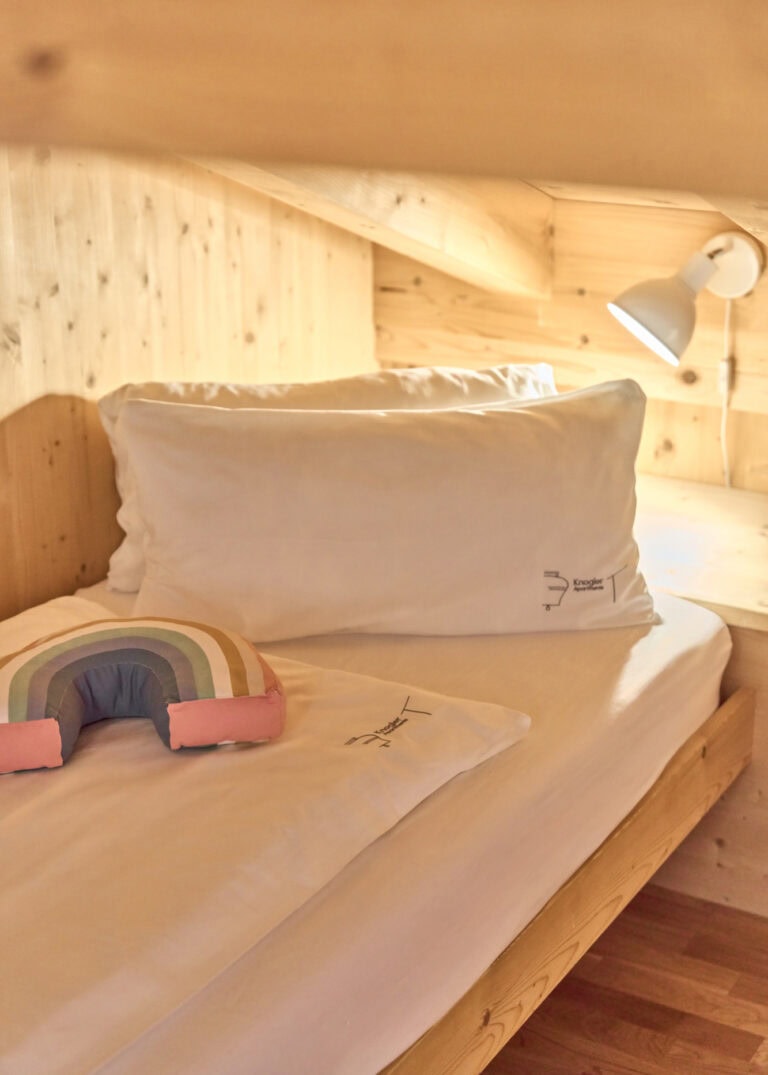 Gemütliches Bett mit Regenbogenkissen im Kinderzimmer der Ferienwohnung Geigelstein.